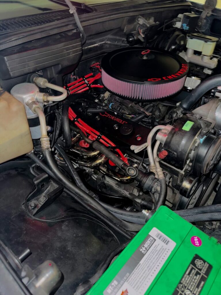 Close-up of a car engine with highlighted red components, including wires and branded cover, and a visible green battery.