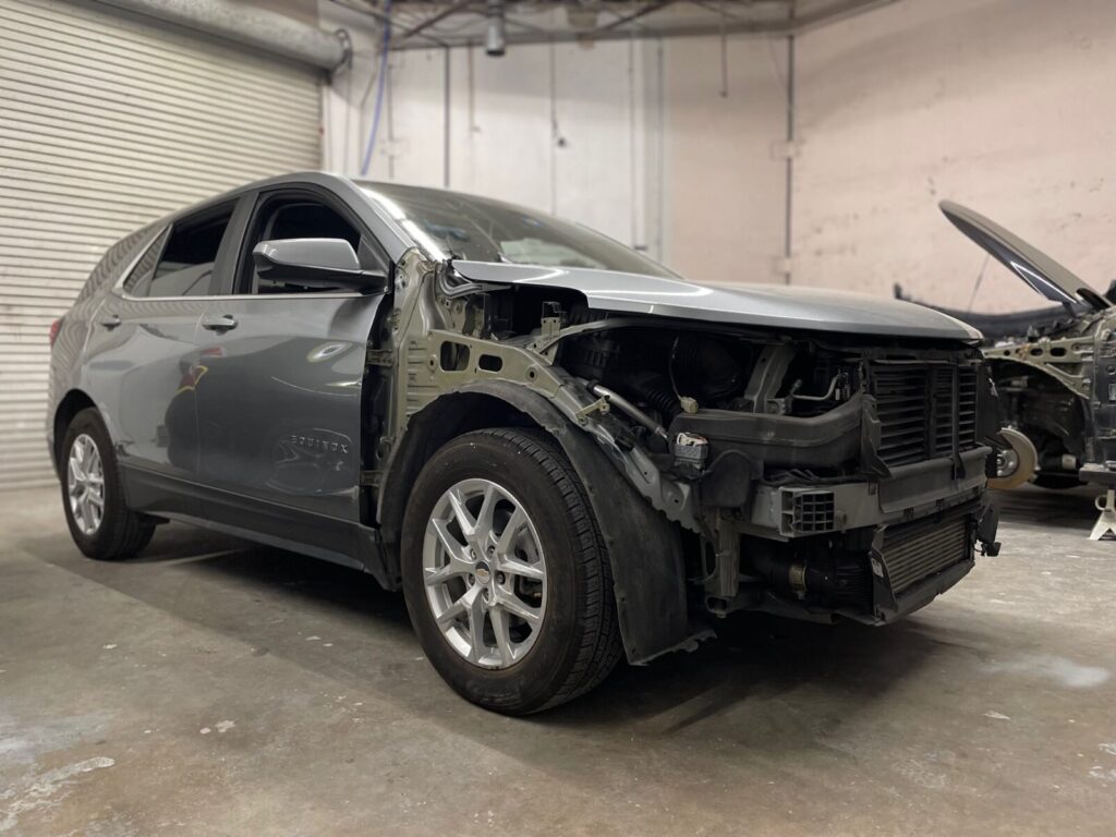 A damaged gray car inside a garage with its front bumper and headlights removed, exposing internal components.